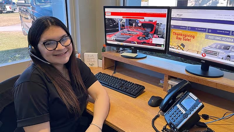 Young woman in glasses and a headset smiling at her desk with two computer monitors displaying car advertisements.