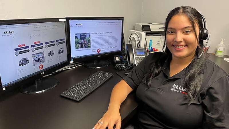 A smiling woman with a headset, wearing a black Kelley Buick GMC shirt, sits at a desk with two computer monitors.