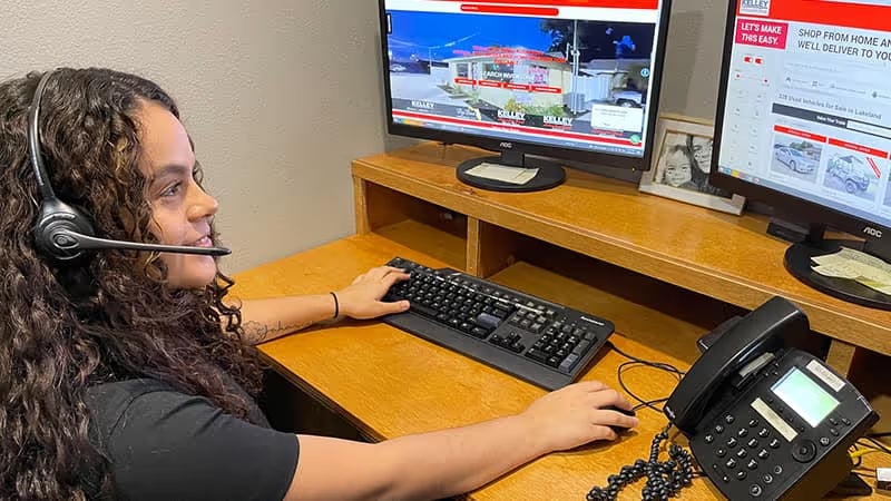 A customer service representative wearing a headset sits at a desk with two computer screens displaying a KELLEY website while she types.