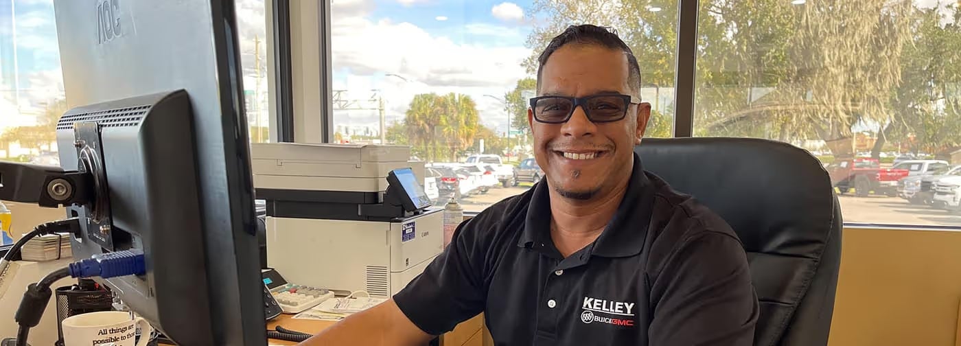 A man wearing glasses and a black Kelley Buick GMC polo shirt smiles from an office desk.