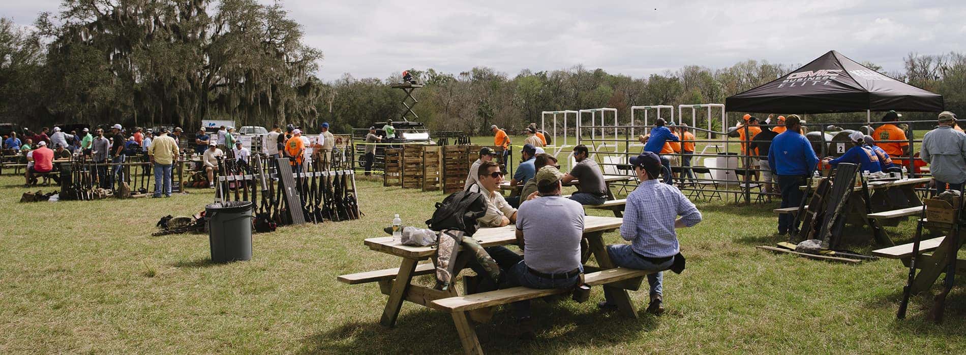 A large group of men gathered outdoors at a shooting range, some seated at picnic tables, others standing by rifle racks.